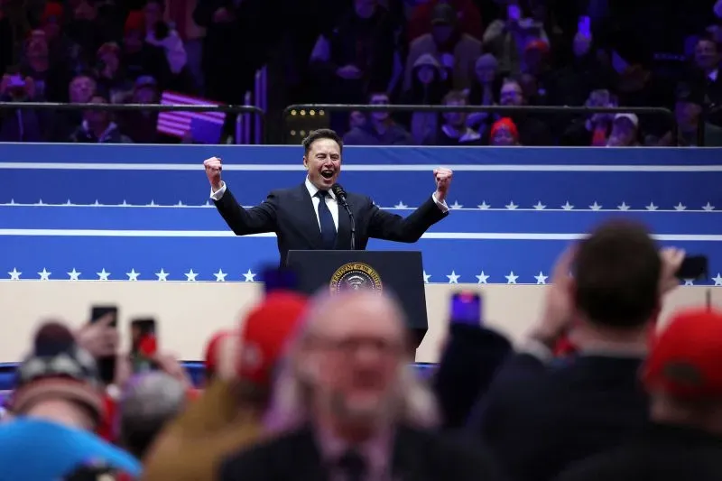 إيلون ماسك يتحدث بحماس على المنصة، محاطاً بجماهير ترتدي قبعات حمراء، مع وجود علم أمريكي خلفه، خلال حدث سياسي.