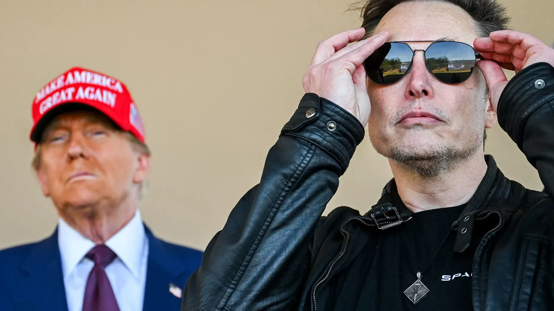 إيلون ماسك يرتدي نظارات شمسية ويضع يديه على رأسه، بينما يظهر خلفه تمثال نصفي لدونالد ترامب مرتديًا قبعة "اجعل أمريكا عظيمة مرة أخرى".