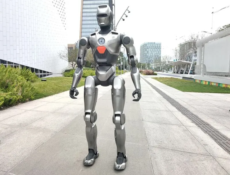 روبوت بشري معدني يقف في منطقة حضرية، مع خلفية تضم مباني حديثة وأشجار، يعكس تقدم الصين في تكنولوجيا الروبوتات.