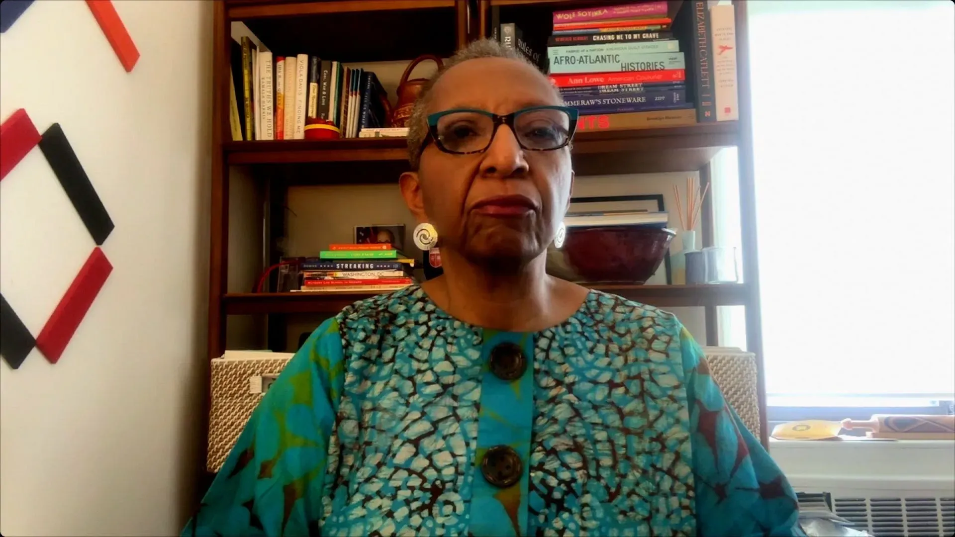 امرأة ترتدي قميصًا مزخرفًا وتضع نظارات، تجلس أمام مكتبة مليئة بالكتب، تعبر عن مخاوفها بشأن تأثير إغلاق المجلس الوطني لعلاقات العمل على حقوق العمال.