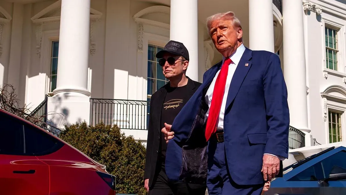 إيلون ماسك ودونالد ترامب يتحدثان خارج البيت الأبيض، مع سيارة تسلا حمراء في المقدمة، مما يعكس العلاقة بين الأعمال والسياسة.