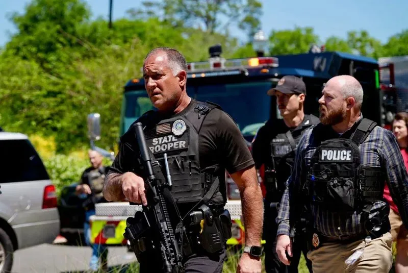 شرطي مسلح يرتدي سترة واقية، يتقدم نحو موقع حادث إطلاق نار، بينما تتواجد سيارات الطوارئ في الخلفية.