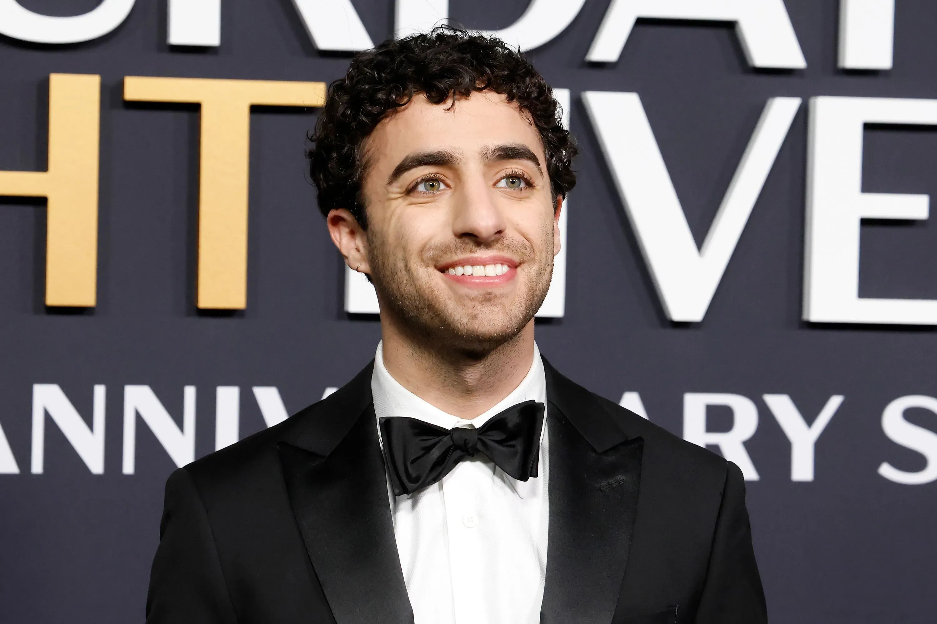 إميل واكيم مبتسم يرتدي بدلة رسمية، يظهر خلفه شعار برنامج "ساترداي نايت لايف"، معبرًا عن مشاعره تجاه مغادرته للبرنامج.