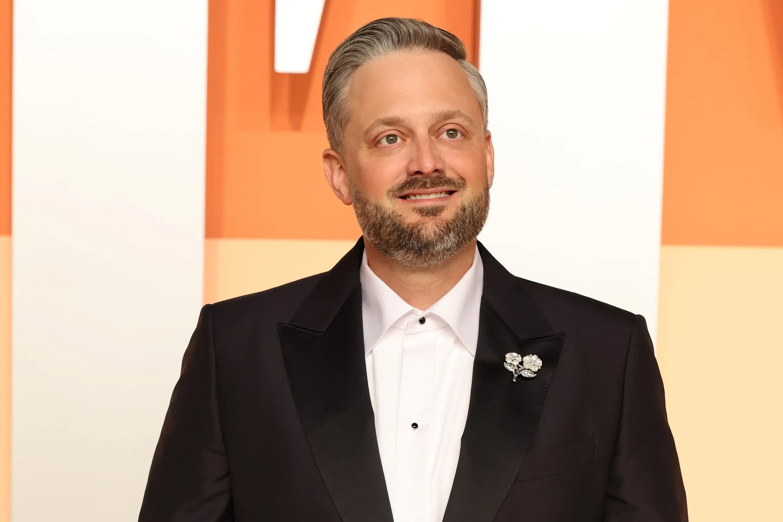 نيت بارجاتزي مبتسم في بدلة رسمية، يستعد لتقديم حفل جوائز إيمي، مع التركيز على الكوميديا النظيفة والترفيه للجمهور.