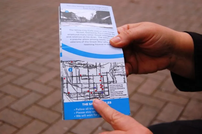 يدان تشير إلى كتيب يحتوي خريطة مواقع المطاعم اليونانية التي تعرضت لأعمال شغب في شارع Yonge بمدينة تورونتو عام 1918، مع شرح تاريخي للأحداث.