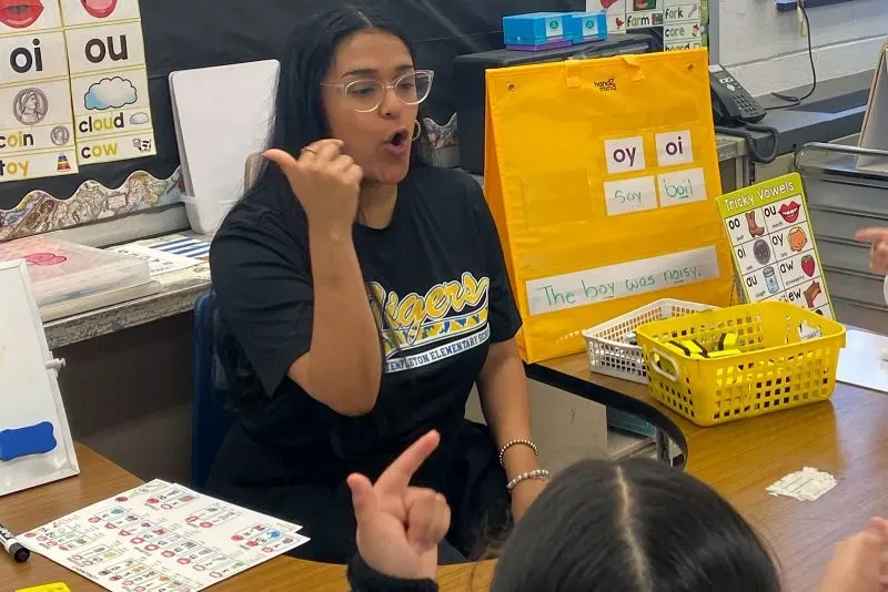 معلمة في فصل دراسي تتفاعل مع طلابها، مستخدمة لغة الإشارة لتعليمهم، بينما تظهر مواد تعليمية على الطاولة.