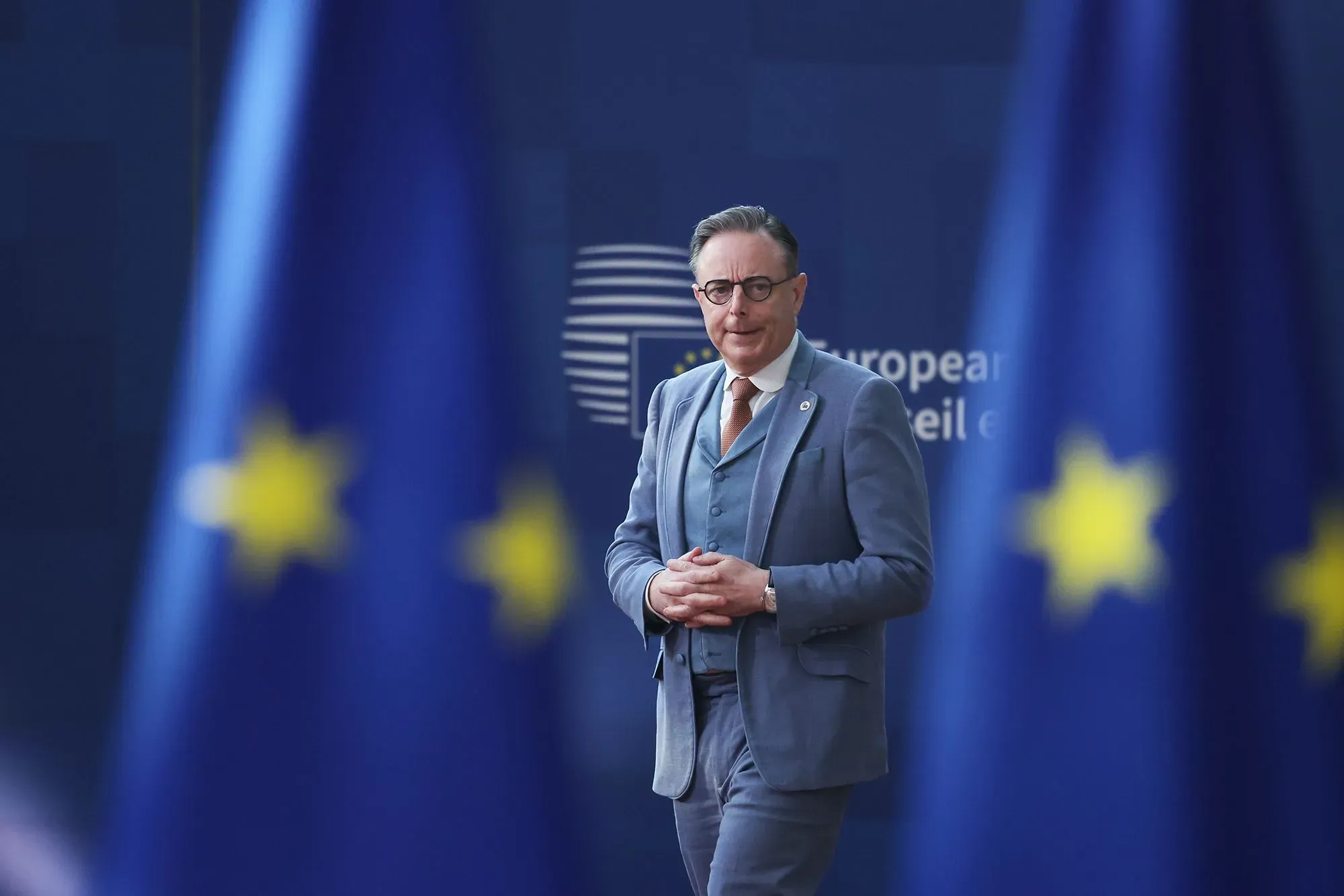 رجل يرتدي بدلة رسمية يقف أمام لافتة تحمل شعار الاتحاد الأوروبي، مع أعلام الاتحاد في الخلفية، في سياق مناقشات حول سياسة الطاقة.