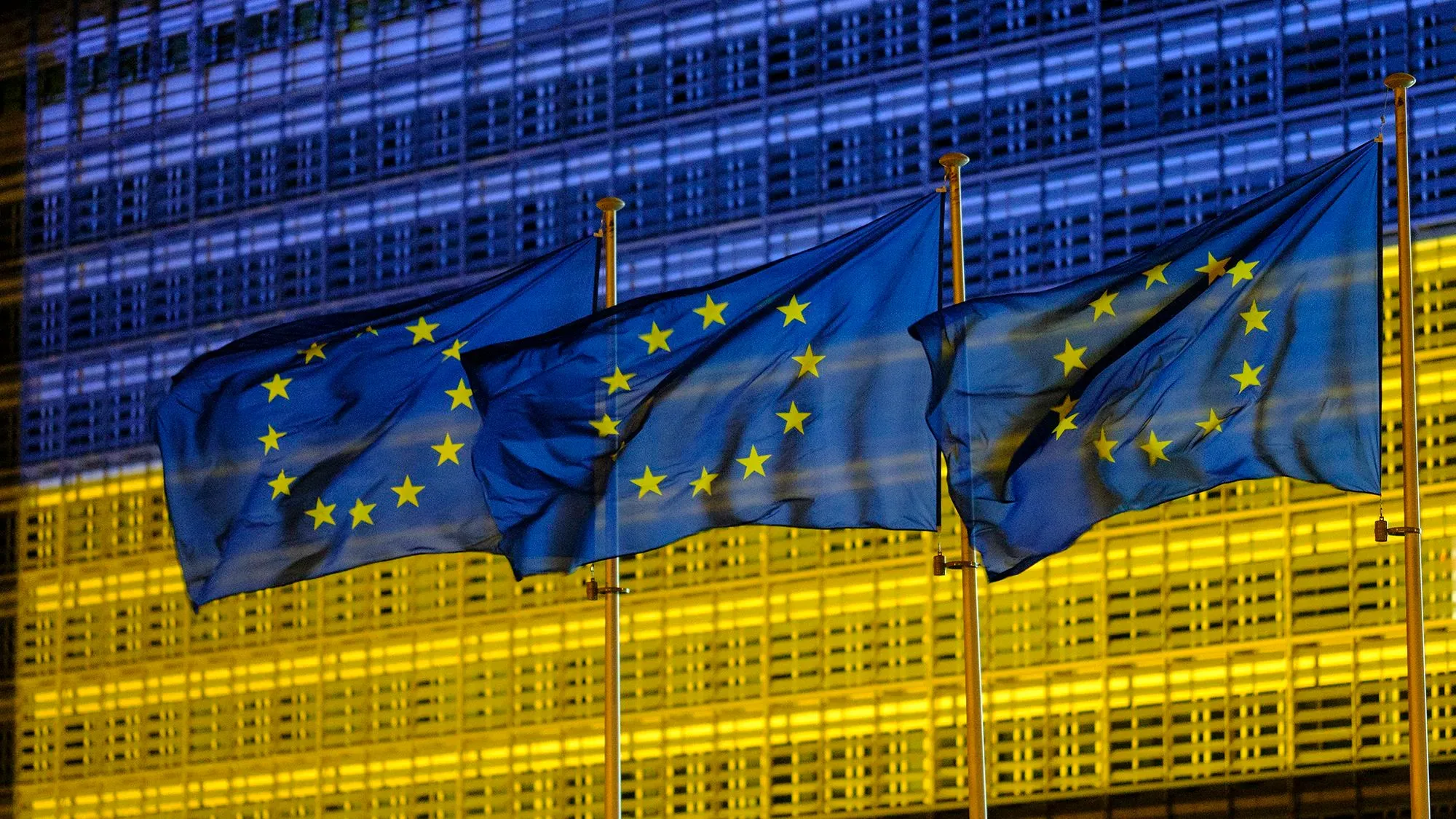 أعلام الاتحاد الأوروبي ترفرف أمام مبنى مضاء بالأزرق والأصفر، مما يرمز إلى الدعم الأوروبي لأوكرانيا في ظل الأزمات الحالية.