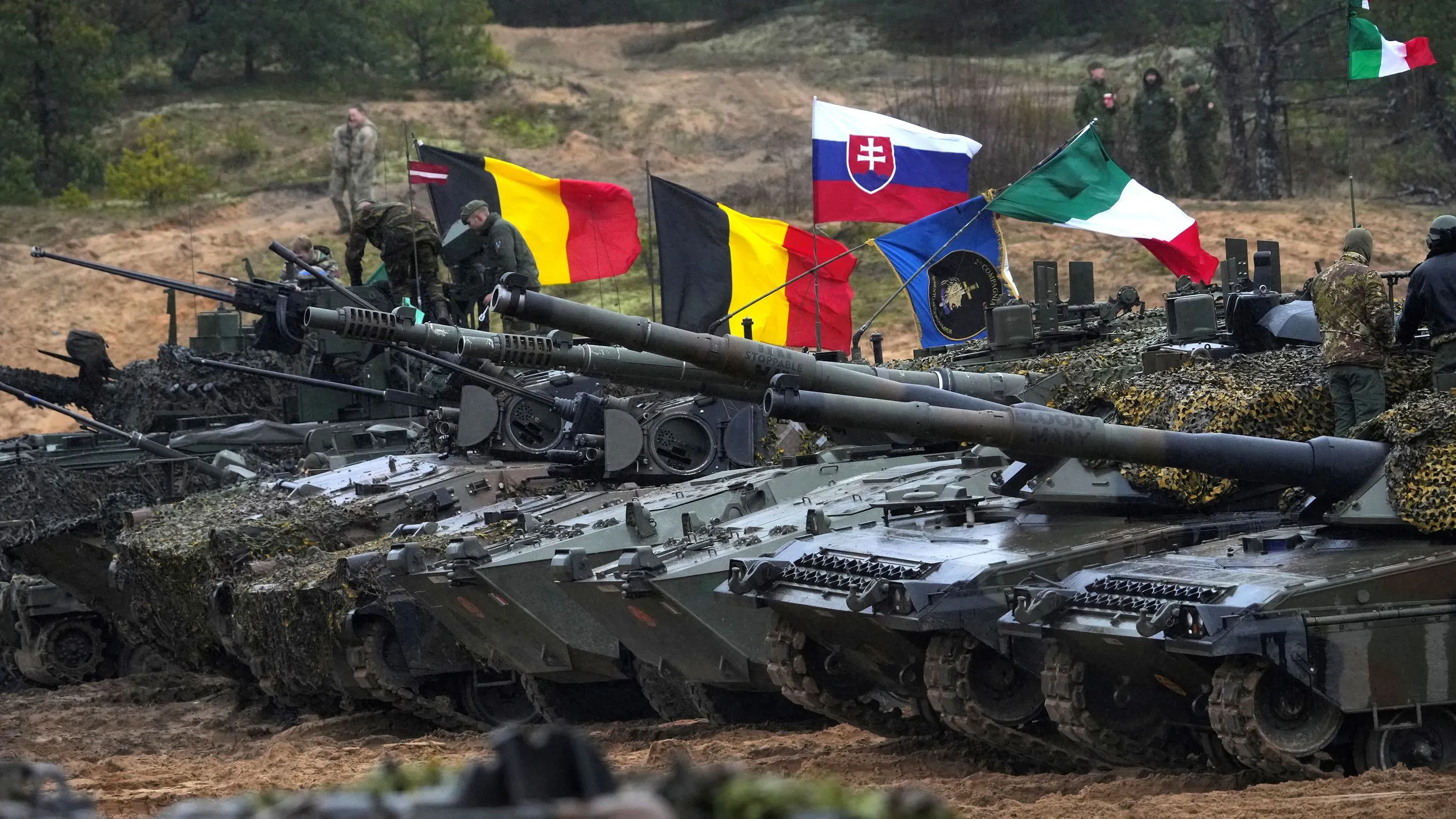 دبابات عسكرية متراصة في عرض عسكري، مع أعلام دول أوروبية مثل لاتفيا وبلجيكا وإيطاليا، تعكس الاستعدادات الدفاعية في سياق التوترات مع روسيا.