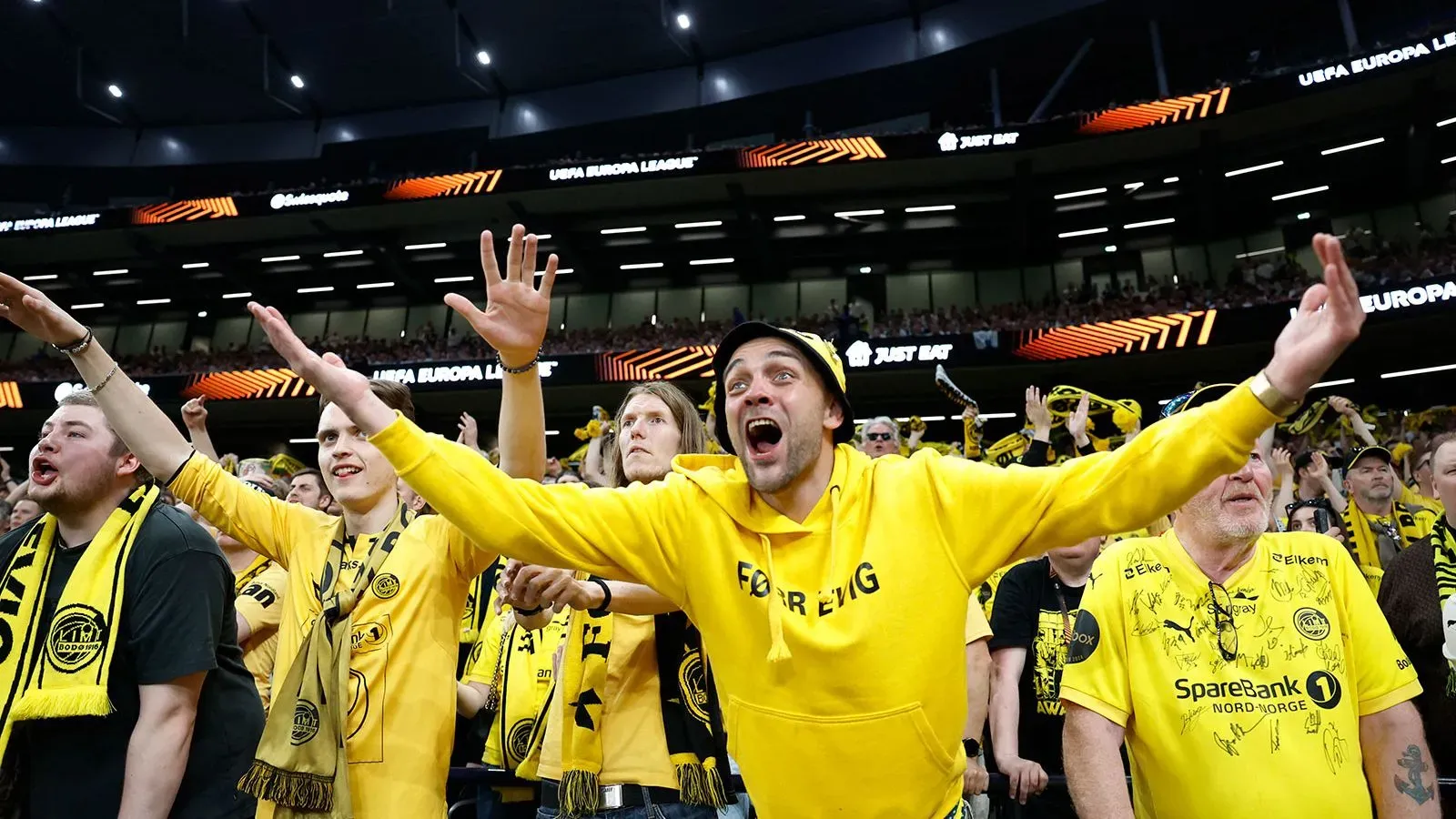 مشجعو فريق بودو/غليمت يرتدون قمصان صفراء ويحتفلون في ملعب توتنهام، مع تفاعل حماسي خلال مباراة نصف نهائي الدوري الأوروبي.