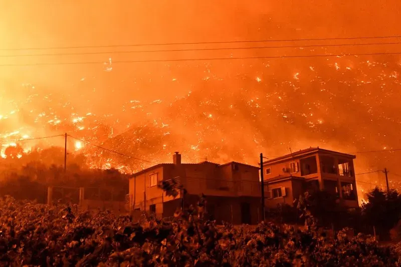 حرائق الغابات تشتعل في الأفق فوق منازل قريبة، مما يخلق مشهدًا مروعًا من الدخان والنيران في سماء حمراء، مما يعكس تأثيرات تغير المناخ على أثينا.