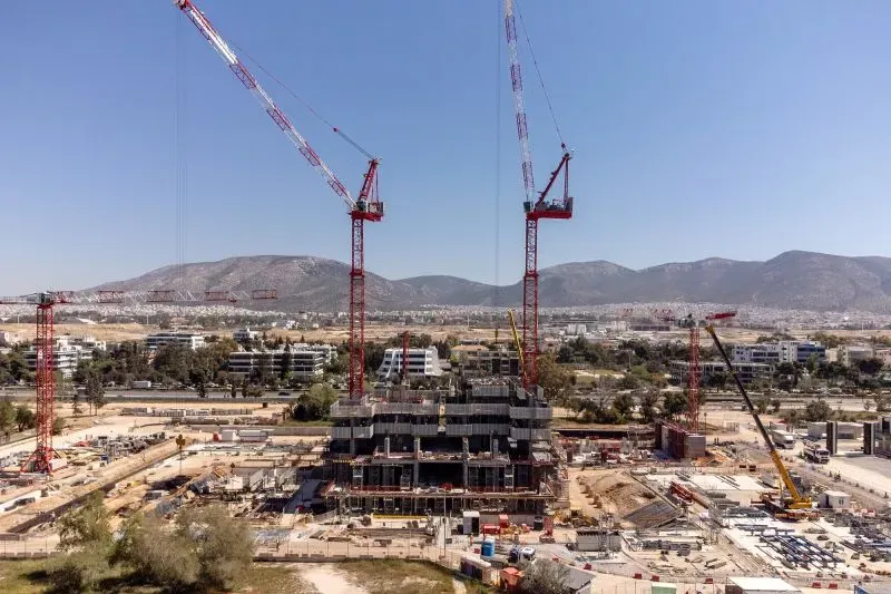 مشهد لموقع بناء في أثينا مع رافعات ضخمة، يظهر التوسع العمراني في المدينة وسط الجبال. تشير الصورة إلى التحديات المناخية والضغط السياحي.