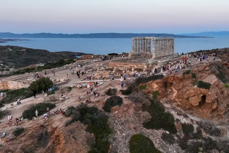 مشهد بانورامي لموقع أثري في اليونان، حيث يتجمع الزوار حول معبد قديم على تلة مرتفعة، مع خلفية بحرية وجبال.
