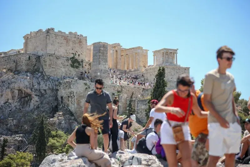 تجمع الصورة زوار الأكروبوليس في أثينا، حيث يتسلقون الموقع التاريخي تحت أشعة الشمس الحارقة، مع ظهور أعمدة البارثينون في الخلفية.