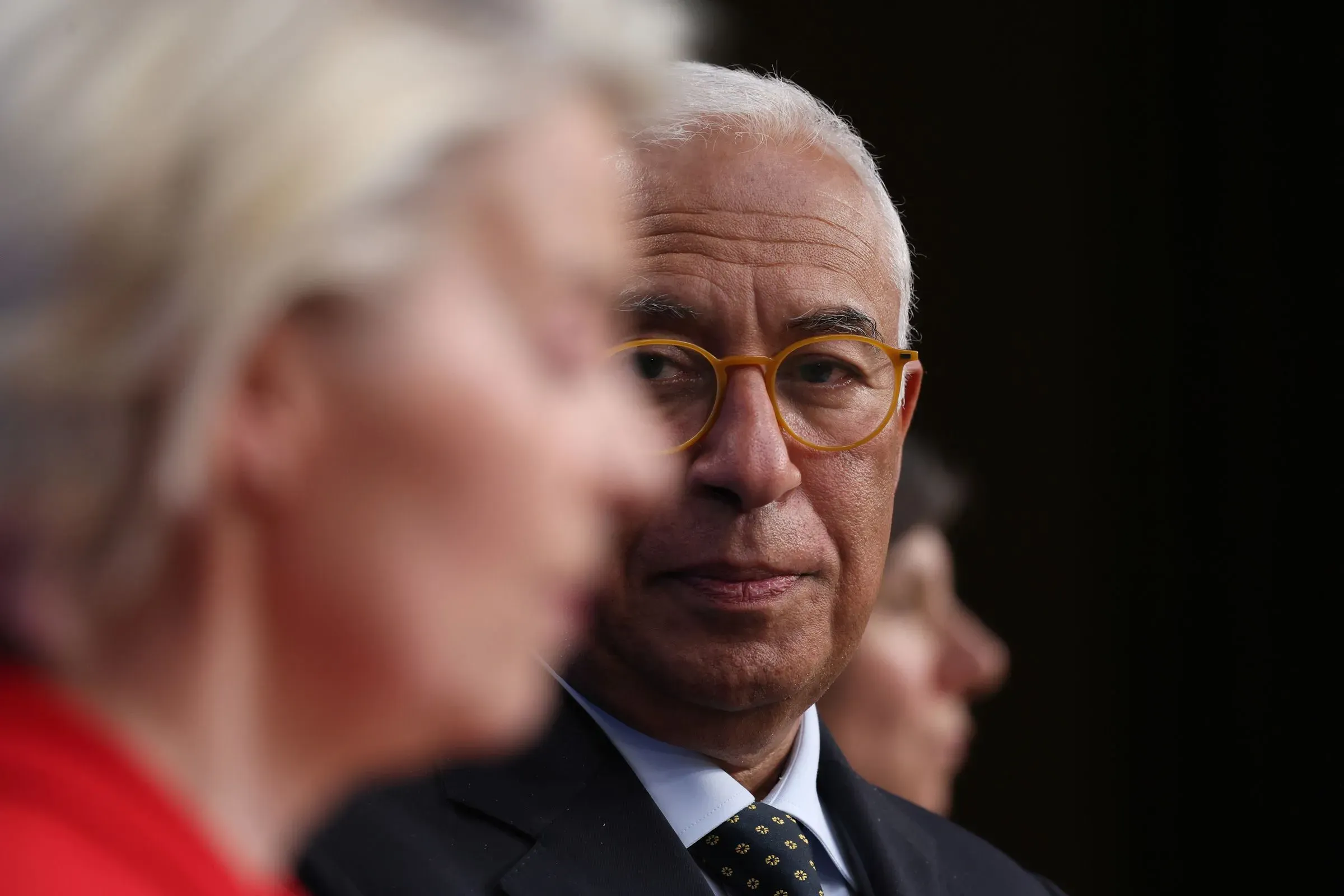 صورة لرجل يرتدي نظارات دائرية، يبدو عميق التفكير، بينما تظهر امرأة في الخلفية تتحدث. تعكس الصورة التوترات السياسية الحالية.