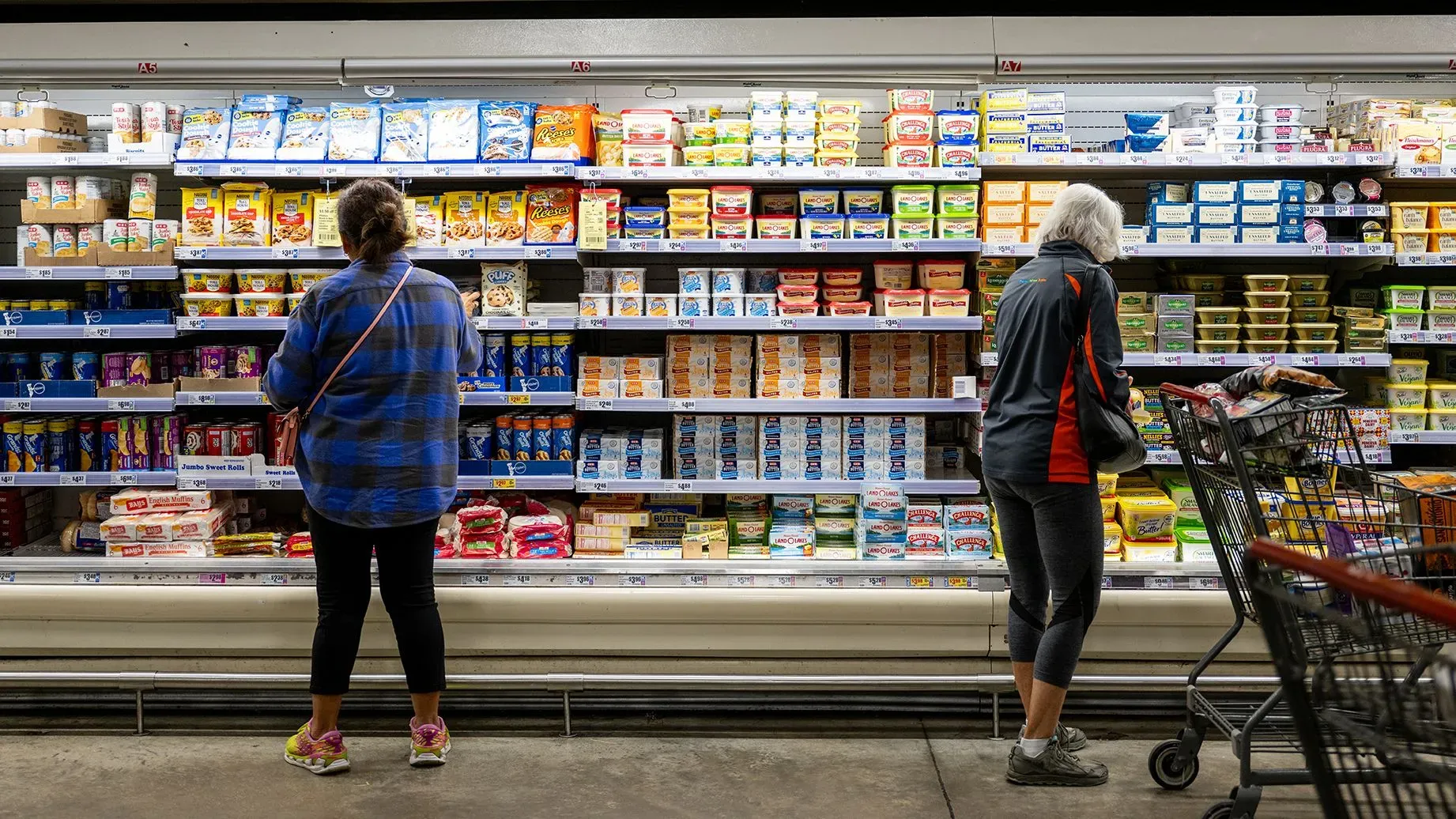 امرأتان تتسوقان في متجر، تقف إحداهما أمام رفوف مليئة بمنتجات الألبان والمأكولات، مما يعكس حالة السوق والاقتصاد الحالي.