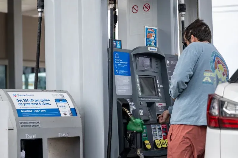 رجل يستخدم محطة وقود لملء سيارته بالبنزين، مع وجود لافتات توضح أسعار الوقود والعروض المتاحة.