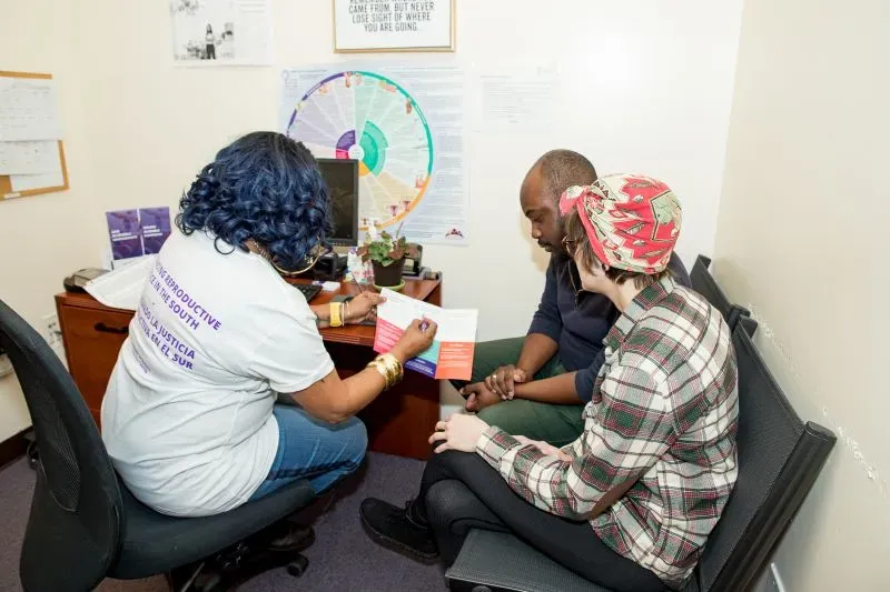 مقدمة استشارة في عيادة للإجهاض، حيث تجلس امرأة ترتدي قميصًا يحمل شعار الرعاية الصحية مع رجل وامرأة أخرى، يتفاعلون مع المستشار.