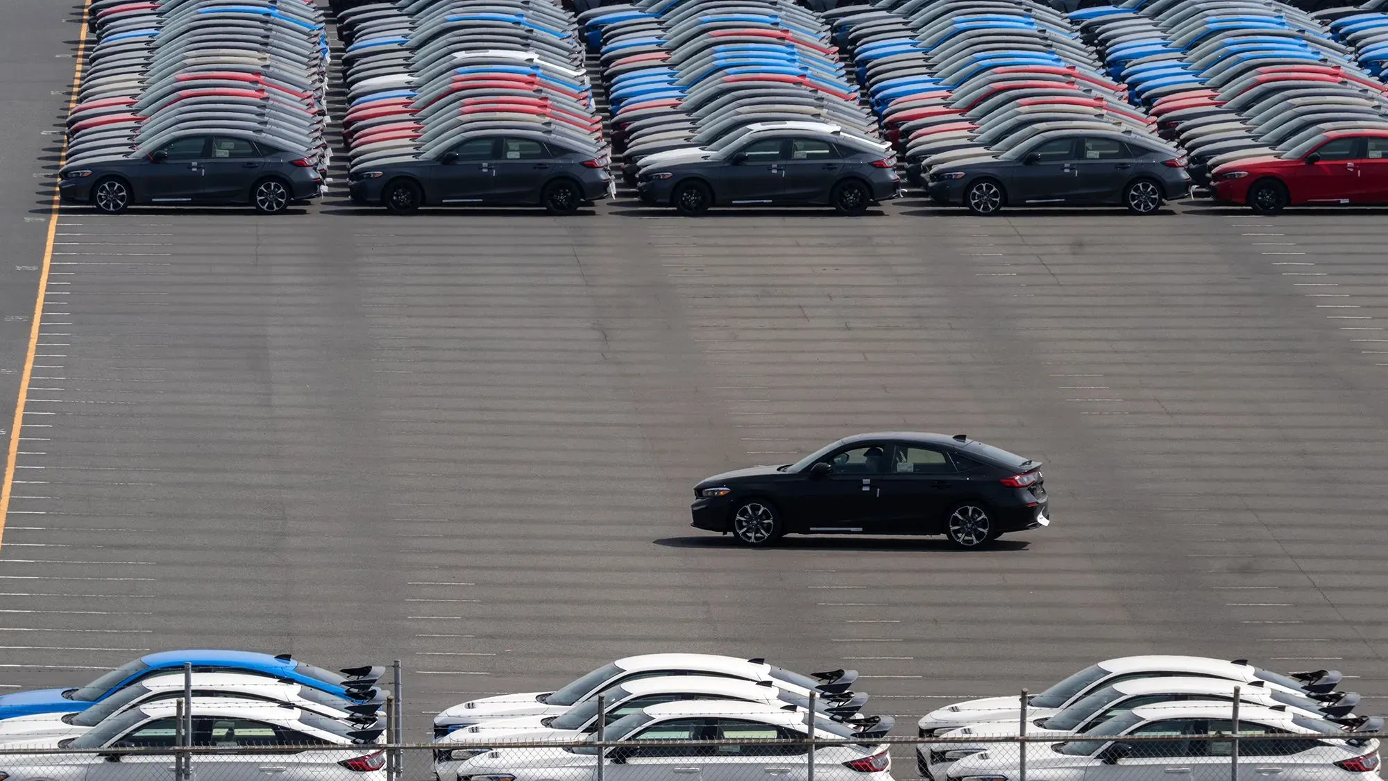 صف من السيارات في ساحة انتظار مزدحمة، مع سيارة سوداء تسير بين صفوف من السيارات بألوان متنوعة، مما يعكس تأثير التعريفات الجمركية على الصناعة.