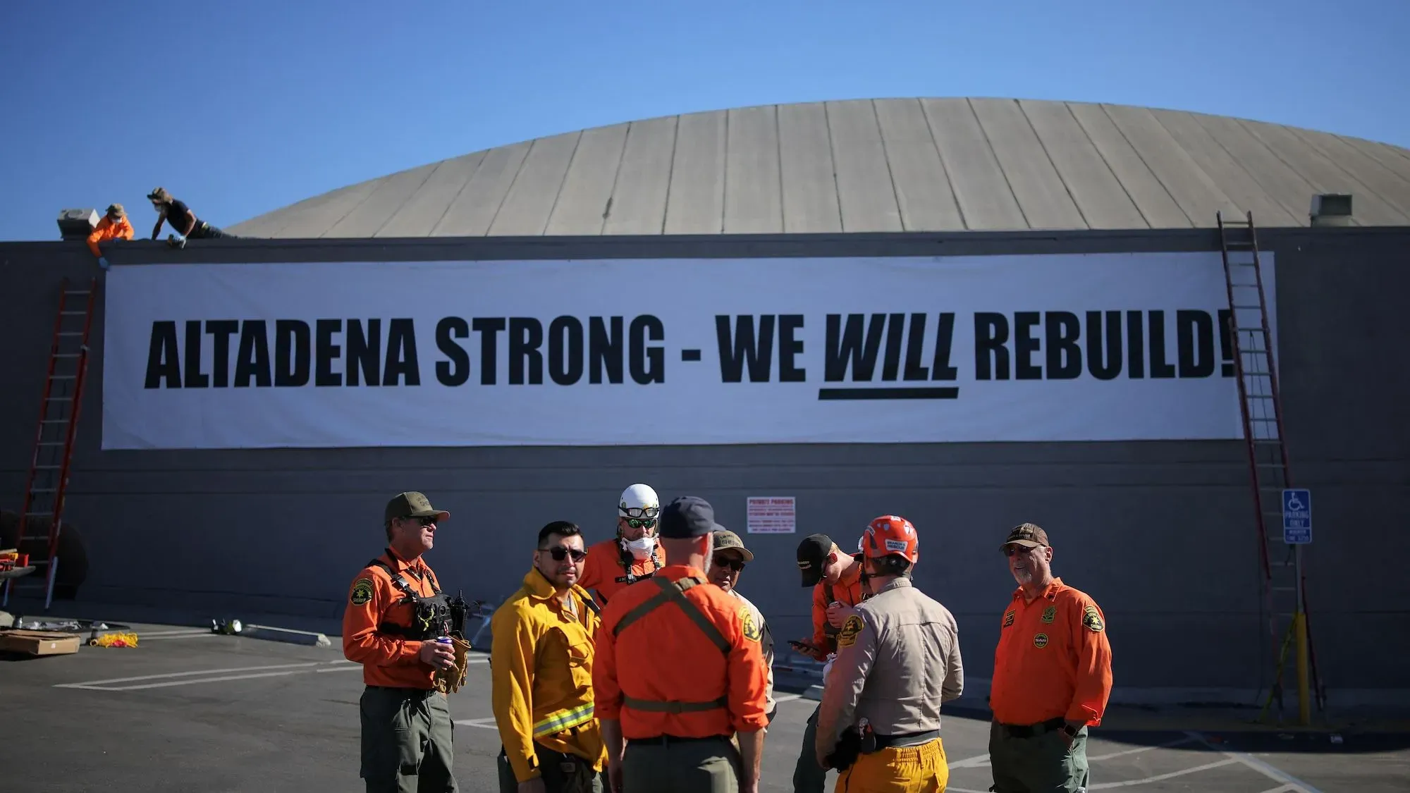 لافتة كبيرة مكتوب عليها "ألتا دينّا قوية - سنعيد البناء!"، مع مجموعة من المتطوعين ورجال الإطفاء في موقع العمل.