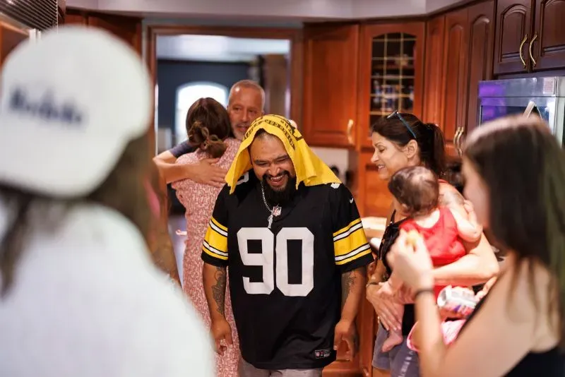 ساشا وخوليو ميندوزا في تجمع عائلي في مطبخ منزلي، حيث يظهر خوليو مرتديًا قميص ستيلرز، محاطًا بأحبائه.