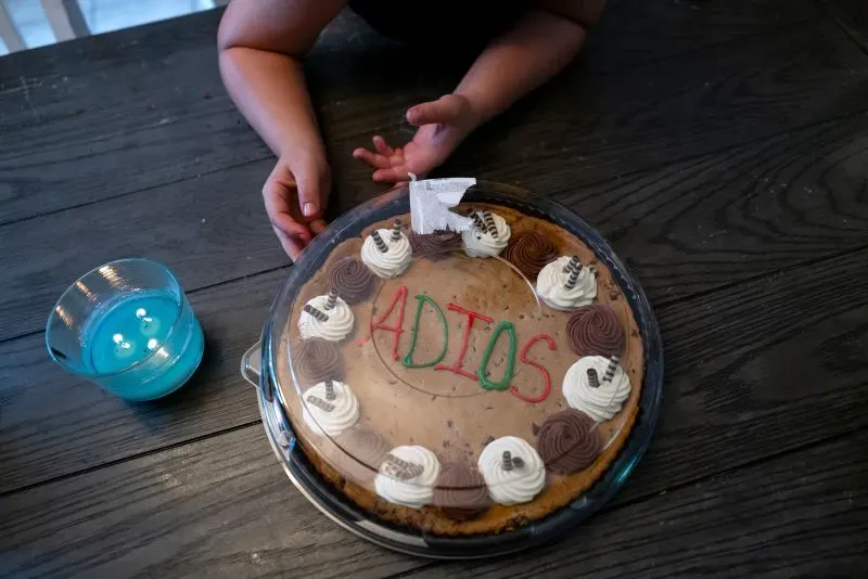 كعكة مكتوب عليها "أديُوس" بألوان زاهية، مع كريمة وشوكولاتة، بينما تظهر يد طفل بالقرب منها، تعبر عن وداع عائلة مهاجرة.