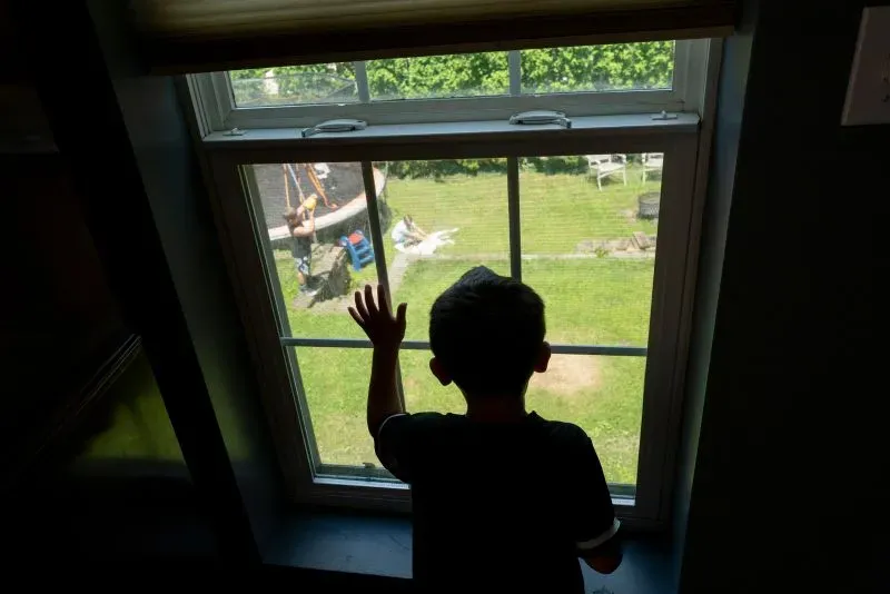 طفل يقف أمام نافذة منزلية، يراقب الحديقة حيث يلعب آخرون، تعبيره يعكس مشاعر الحزن والحنين.