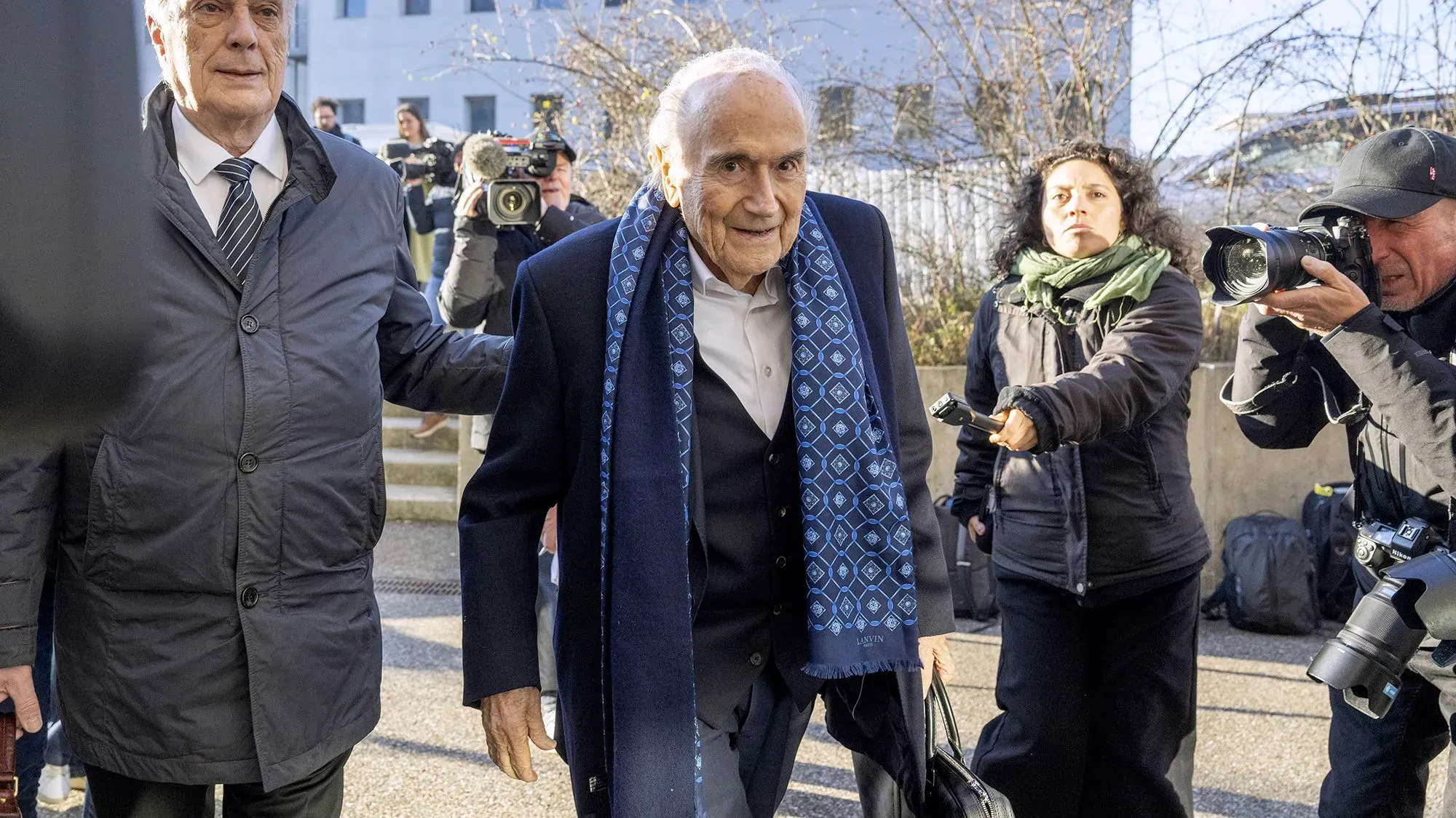 يمثل سيب بلاتر وميشيل بلاتيني أمام المحكمة في سويسرا بتهمة الاحتيال، مع وجود وسائل الإعلام في الخلفية.