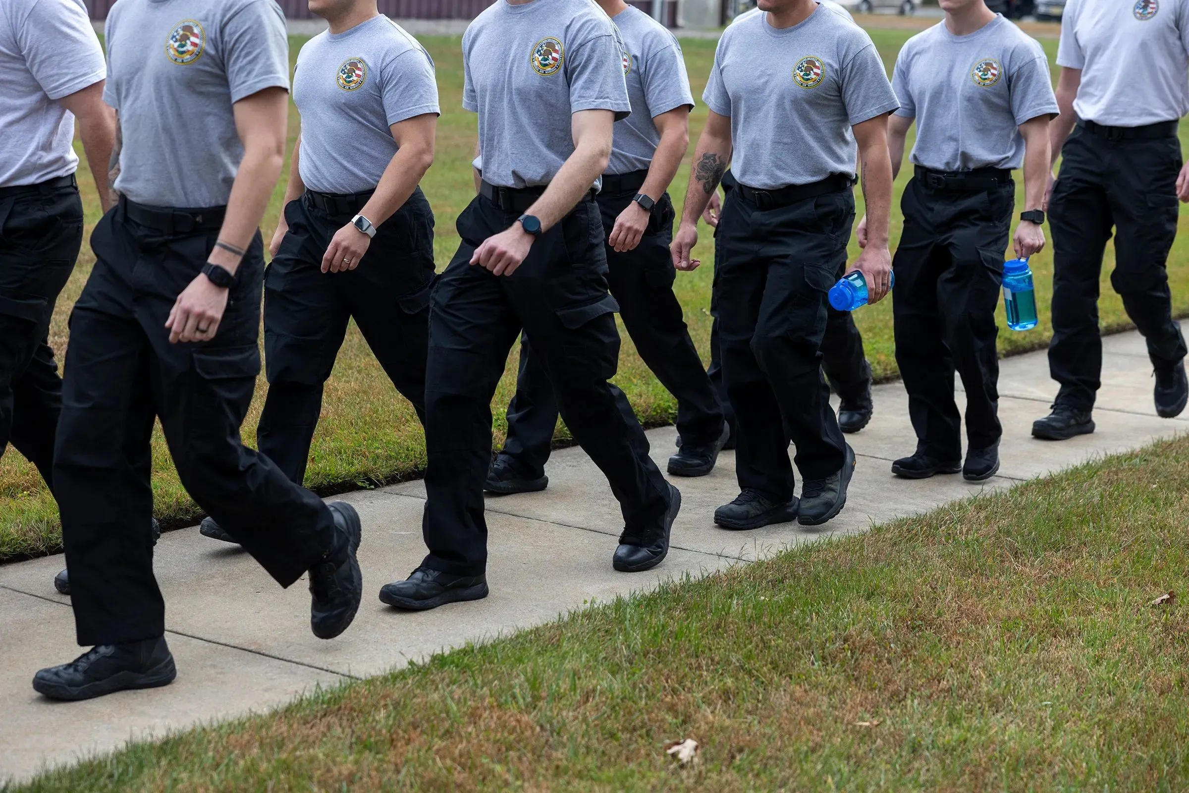 مجموعة من المارشالات الجوية الفيدرالية يرتدون زيًا موحدًا، يسيرون في صف واحد، مما يعكس جهودهم في تأمين رحلات الترحيل.