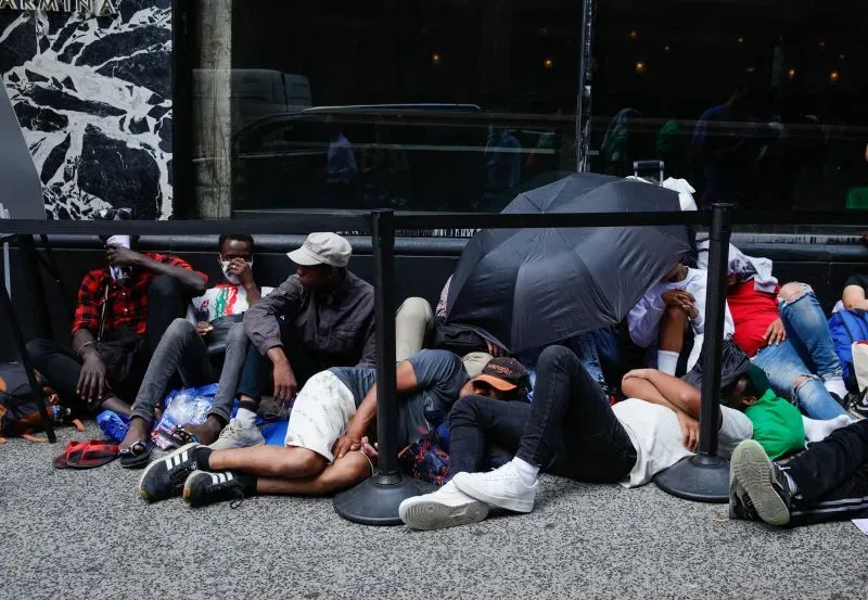 مجموعة من المهاجرين يتجمعون على الرصيف في نيويورك، بعضهم يستخدم مظلة للحماية من الشمس، تعبيرًا عن الظروف الصعبة التي يواجهونها.