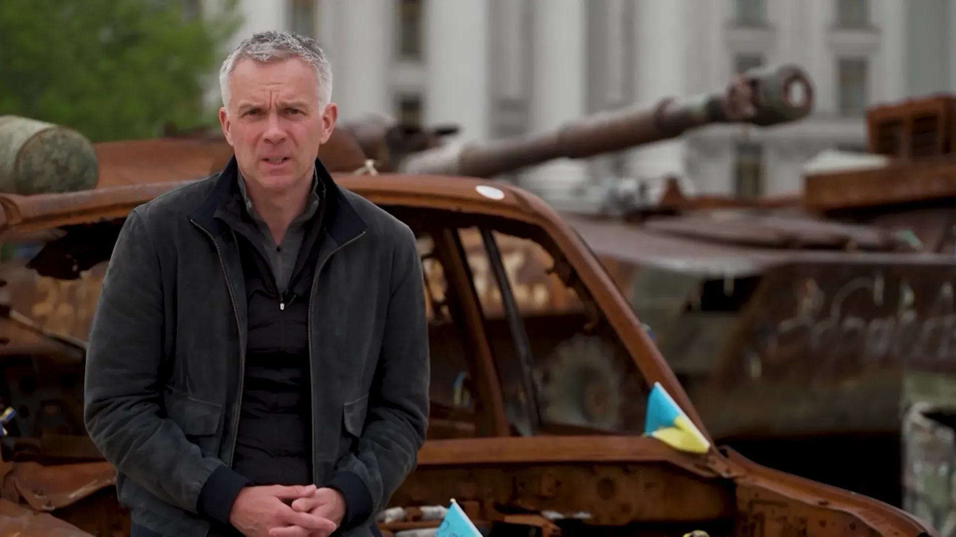 مراسل يقف أمام سيارة مدمرة ودبابة في منطقة متأثرة بالصراع، مع التركيز على الأوضاع الإنسانية في أوكرانيا.