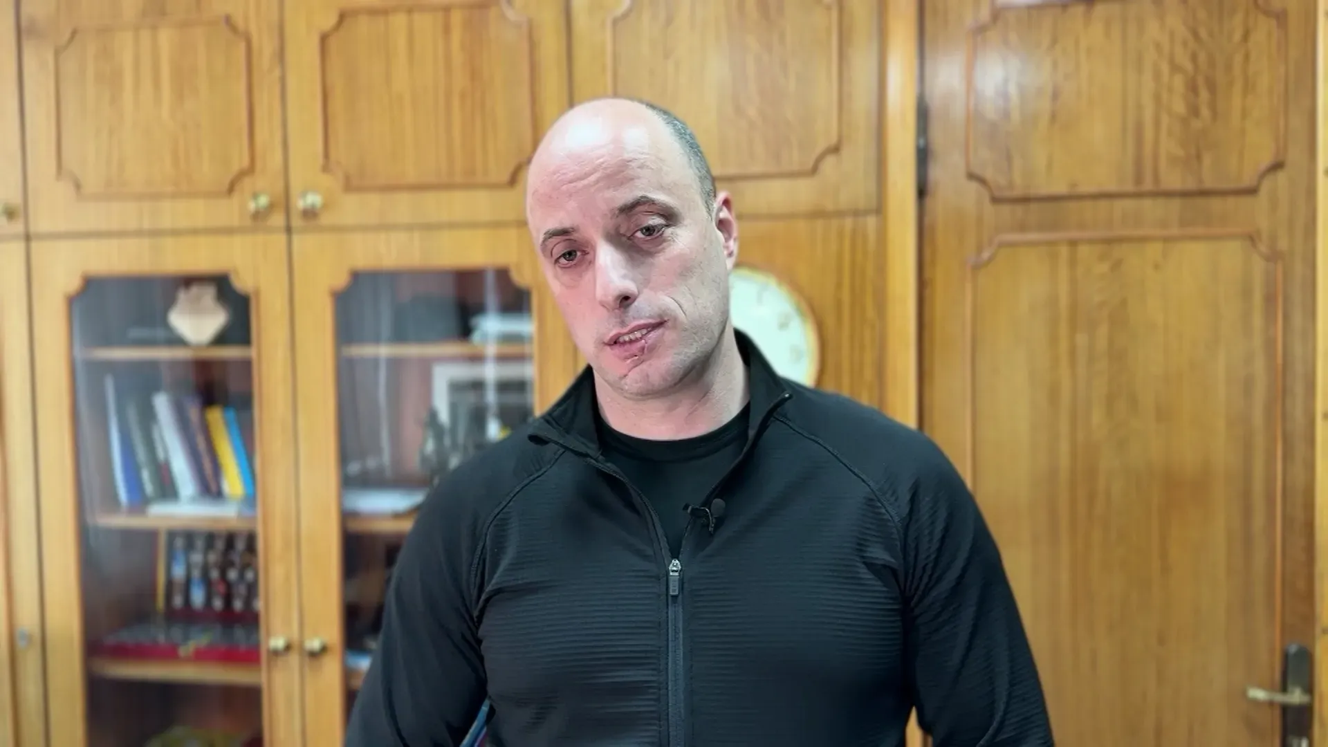 شخص يرتدي ملابس سوداء يتحدث بجدية أمام مكتبة تحتوي على كتب وأغراض أخرى، في سياق تحقيقات حول انتهاكات حقوق الإنسان في أوكرانيا.