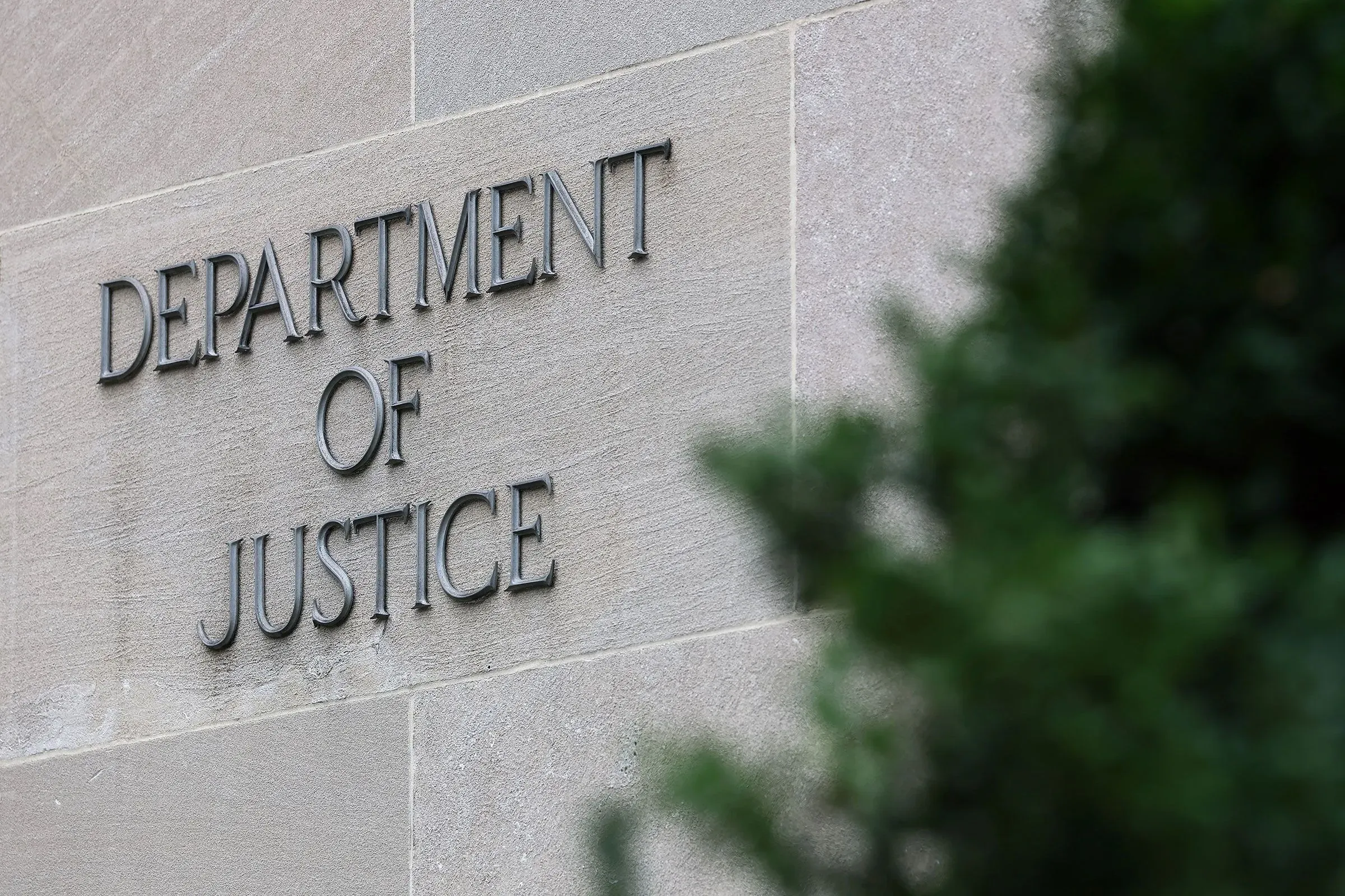 لافتة وزارة العدل الأمريكية مكتوبة على جدار، مع وجود نباتات خضراء في المقدمة، تعكس التغييرات في قيادة المكتب.