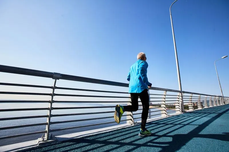 رجل مسن يرتدي ملابس رياضية زرقاء يجري على ممشى بجوار المياه، مما يعكس أهمية النشاط البدني في تحسين البقاء على قيد الحياة لمرضى سرطان القولون.