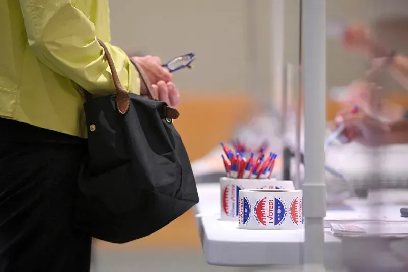 ناخبة ترتدي قميصًا أصفر وتحمل حقيبة سوداء، تقف بالقرب من طاولة الاقتراع التي تحتوي على أدوات التصويت وأقلام.