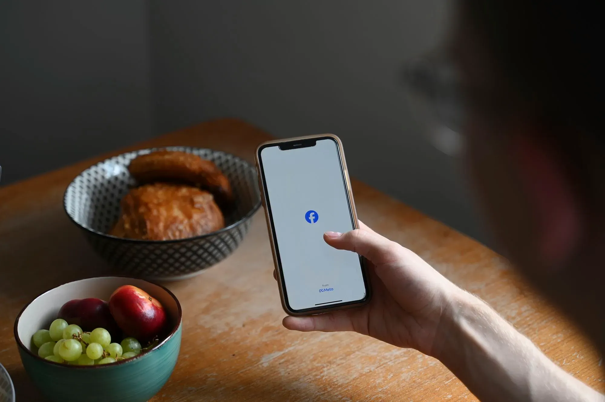 شخص يستخدم هاتفه المحمول لفتح تطبيق فيسبوك، بينما تظهر أطباق من الفواكه والمعجنات على الطاولة، في سياق تسوية متعلقة بخصوصية البيانات.