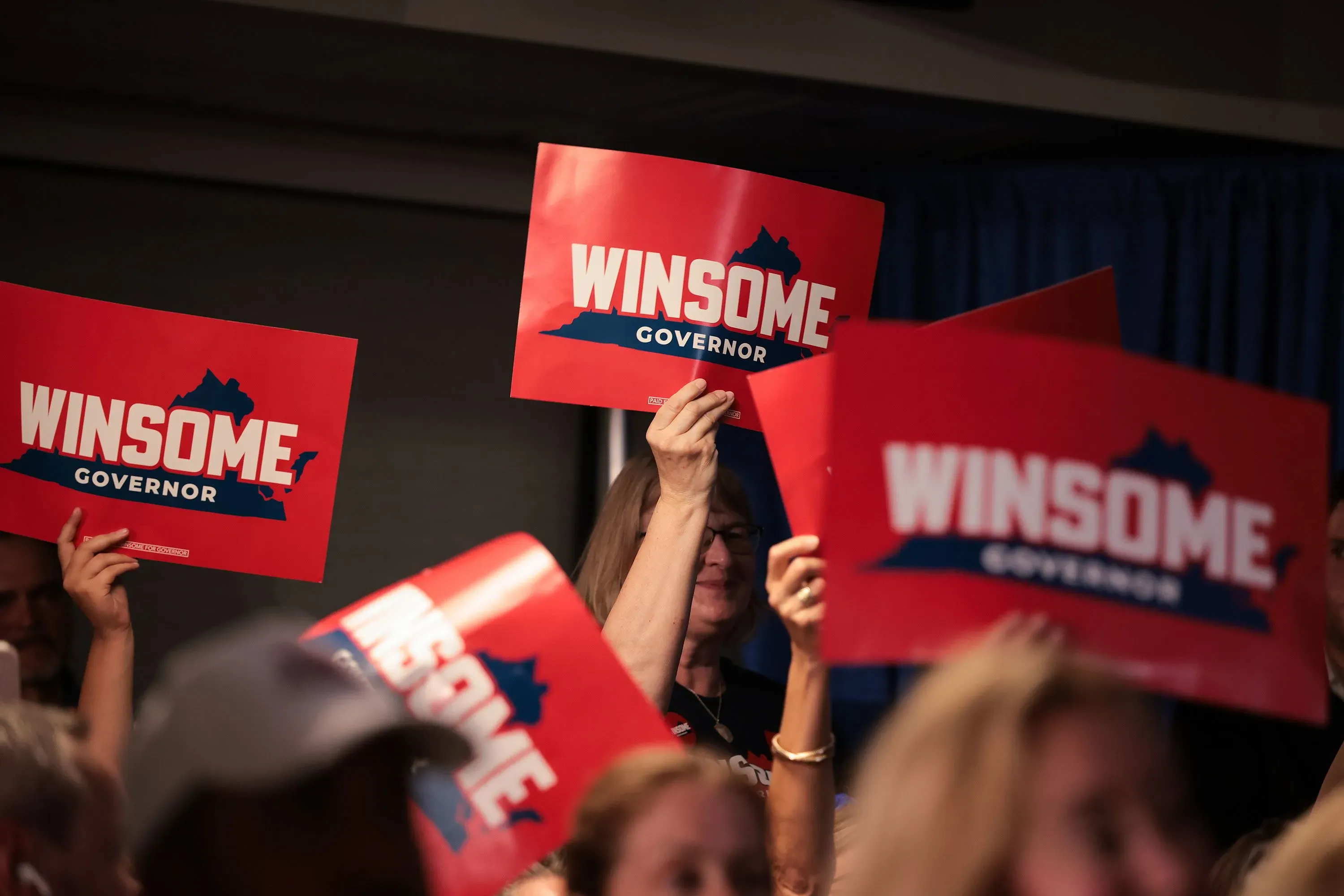 لافتات انتخابية تحمل اسم "وينسوم" مرشحة حاكم ولاية فيرجينيا، تُرفع من قبل مؤيدين في حدث جماهيري.