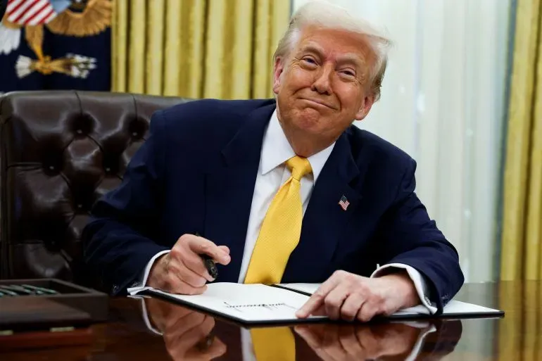 ترامب مبتسم أثناء توقيع وثيقة في المكتب البيضاوي، مع خلفية رسمية تظهر الستائر الذهبية والشعار الرئاسي.