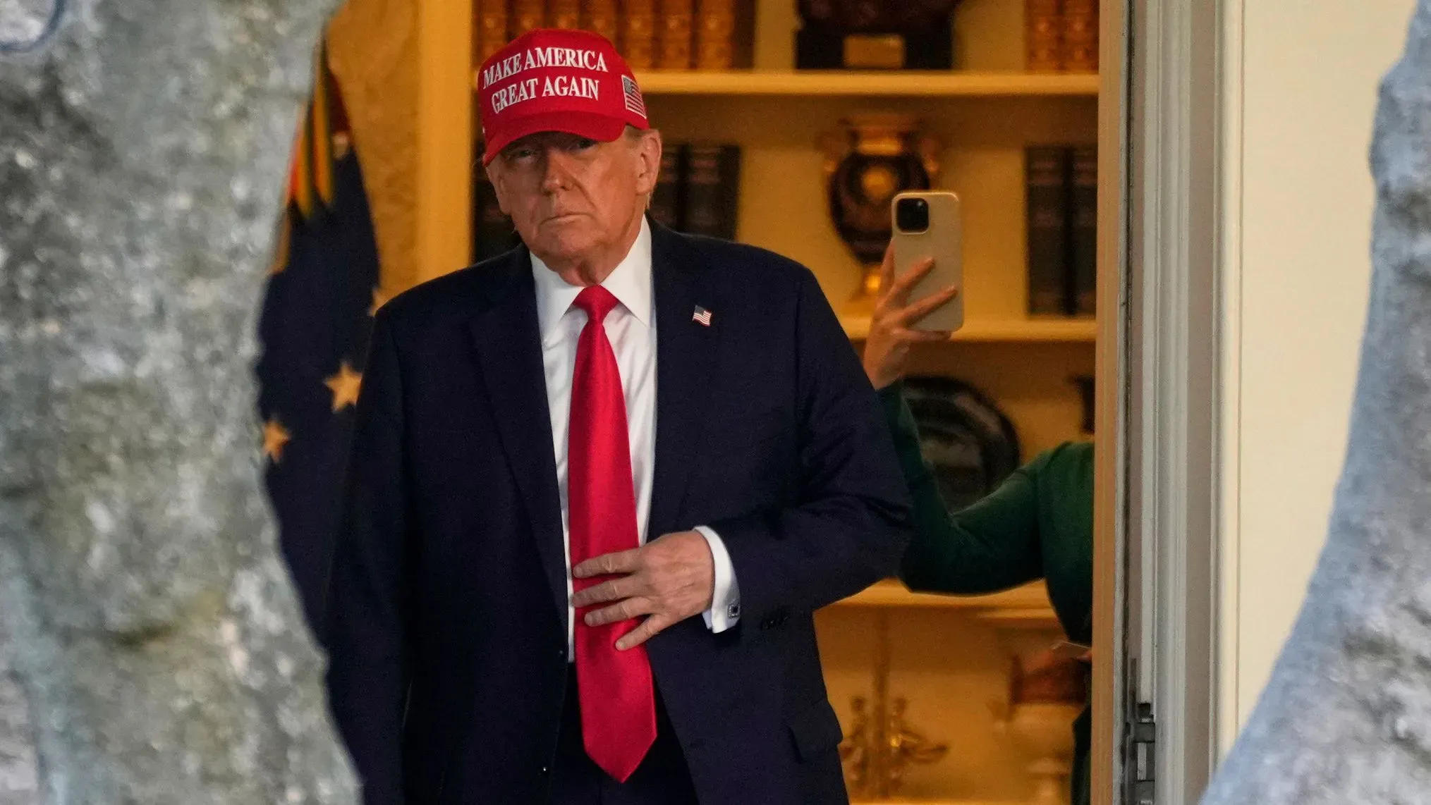 ترامب يرتدي قبعة "اجعل أمريكا عظيمة مرة أخرى" أثناء مغادرته البيت الأبيض، مع وجود شخص آخر في الخلفية يحمل هاتفًا.