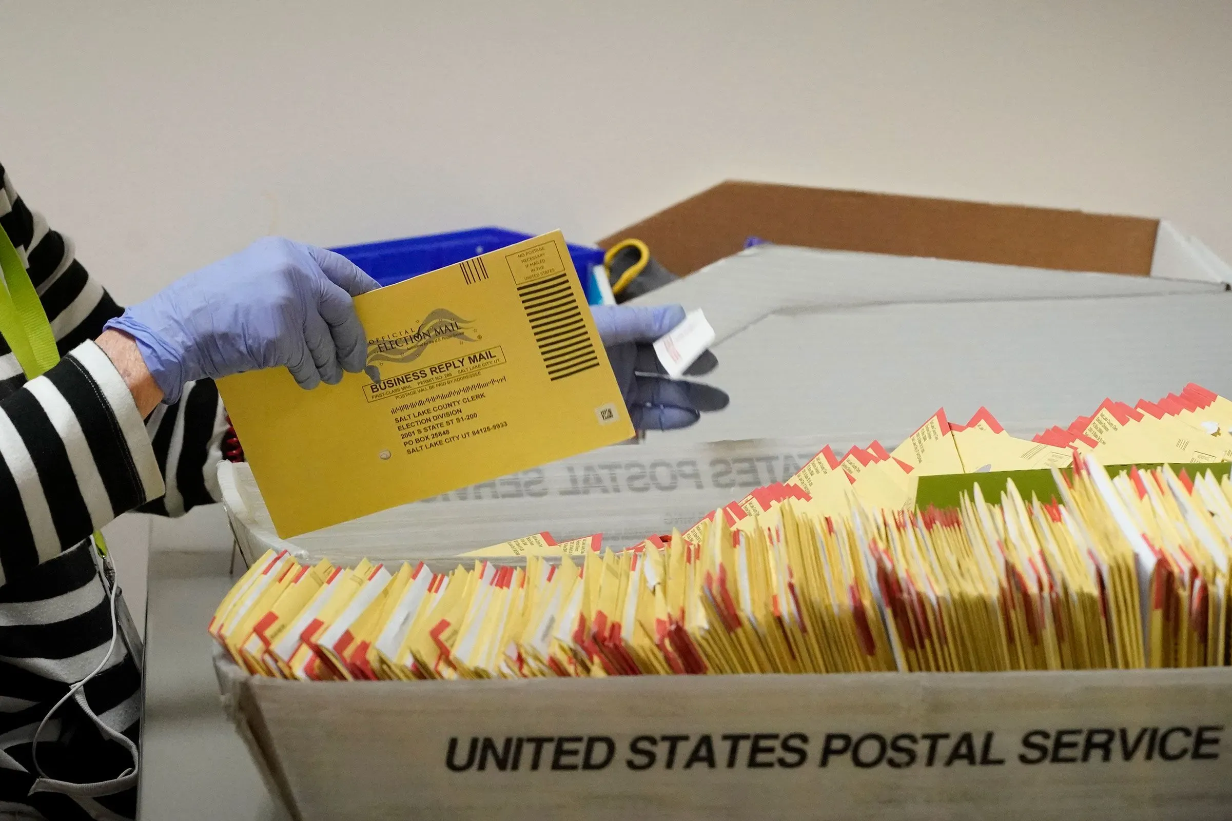 إمرأة ترتدي قفازات تفحص بطاقات اقتراع بريدية صفراء في صندوق مخصص للبريد، في سياق مناقشة حول نزاهة الانتخابات.