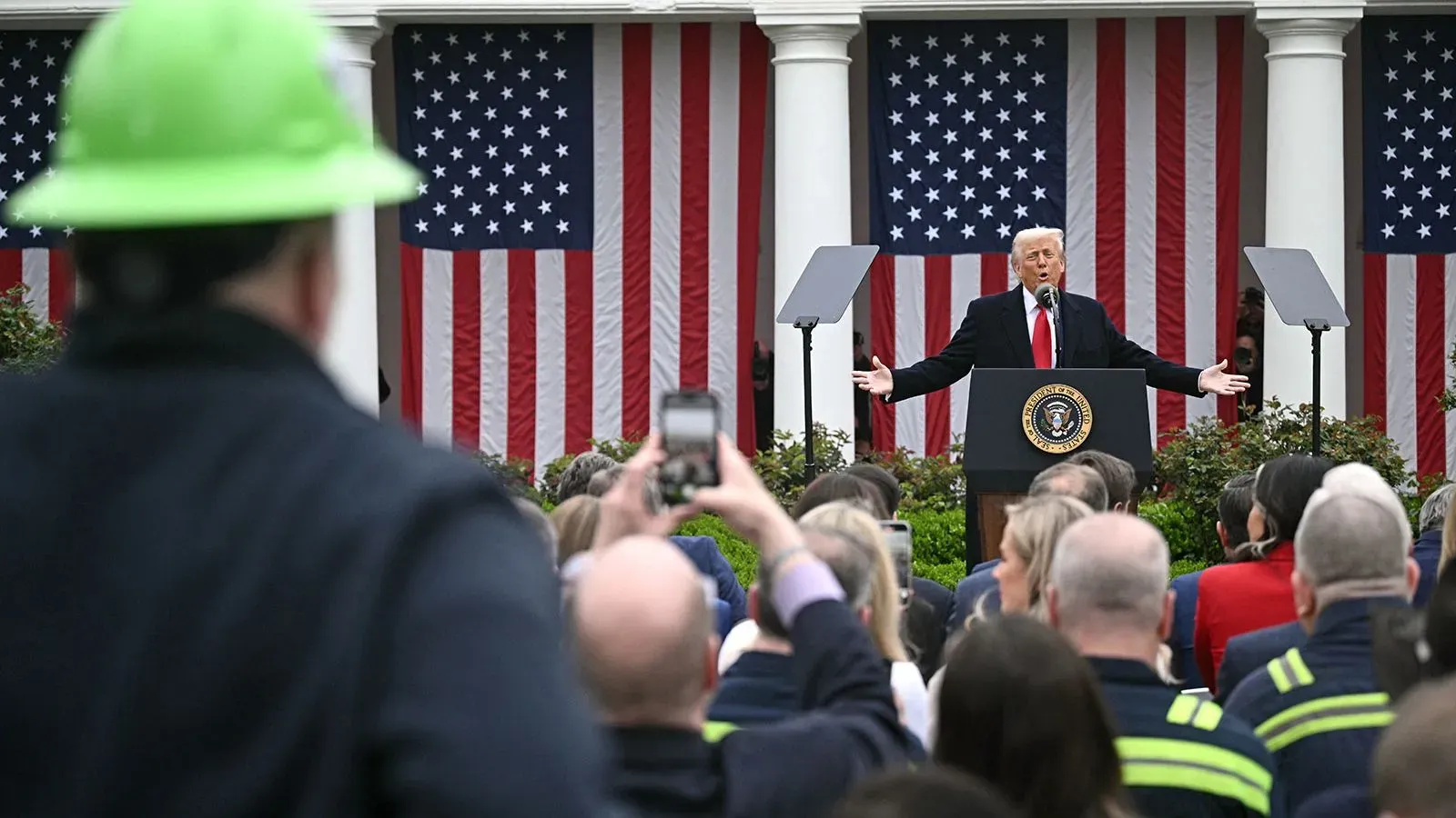 خطاب ترامب في حديقة البيت الأبيض، حيث يتحدث أمام جمهور كبير مع العلم الأمريكي خلفه، مع وجود رجل يرتدي خوذة خضراء في المقدمة.