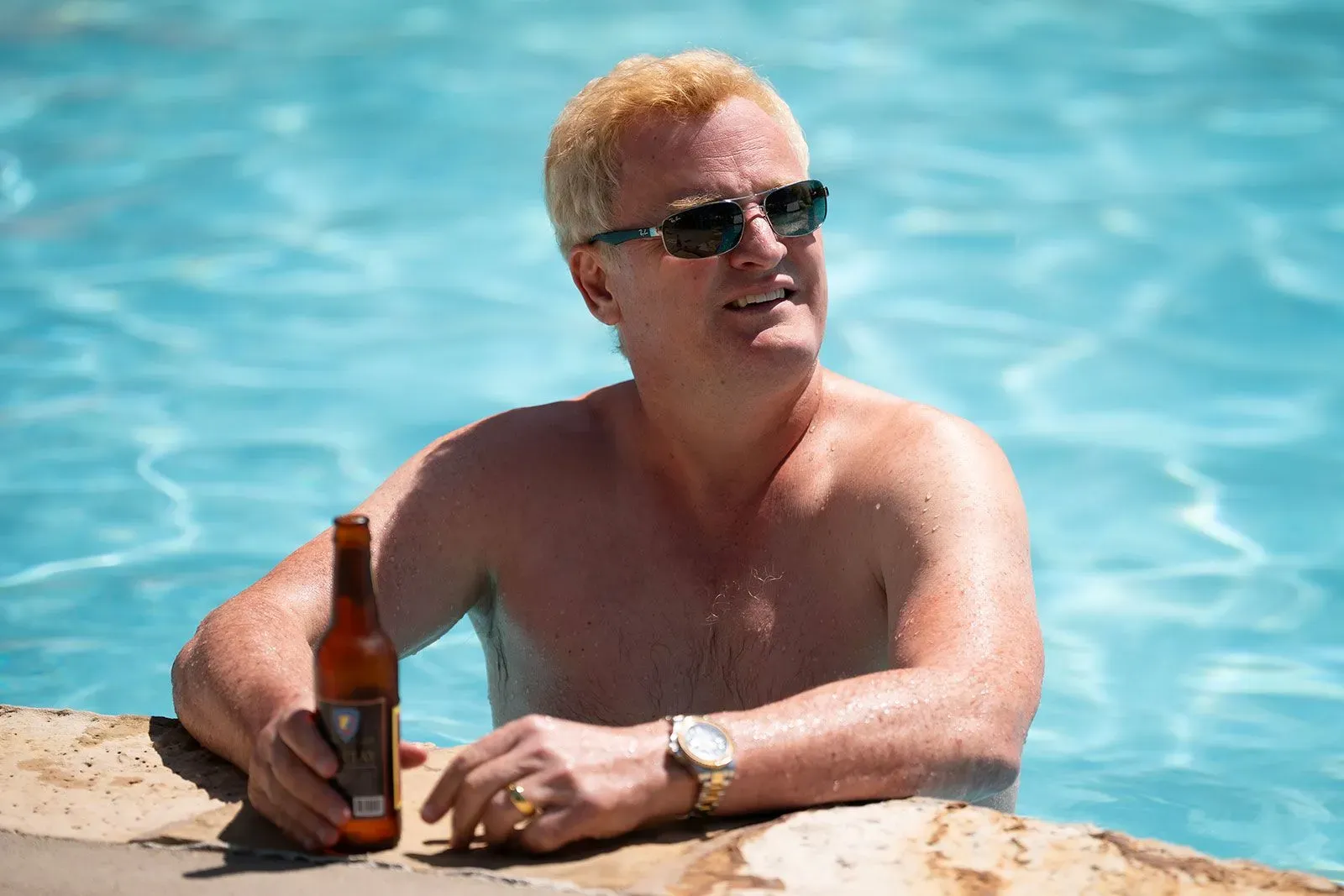 رجل عائم في بركة سباحة، يرتدي نظارات شمسية، ممسكًا بزجاجة مشروب، في مشهد يعكس حياة الرفاهية قبل الأحداث المأساوية لعائلة موردو.