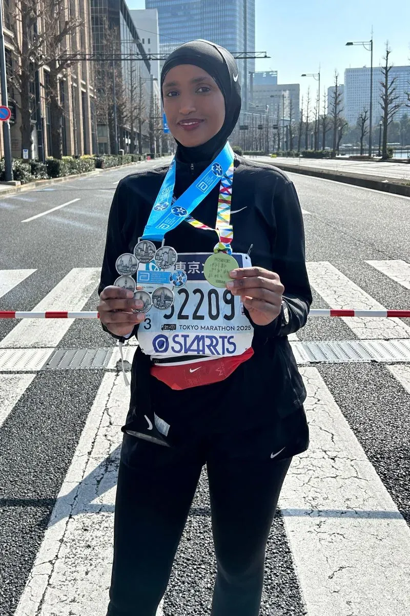 امرأة ترتدي زي الجري الأسود وتحمل ميداليات سباق، تقف في الشارع مع خلفية مباني حضرية، تعبر عن إنجازها في ماراثون طوكيو.