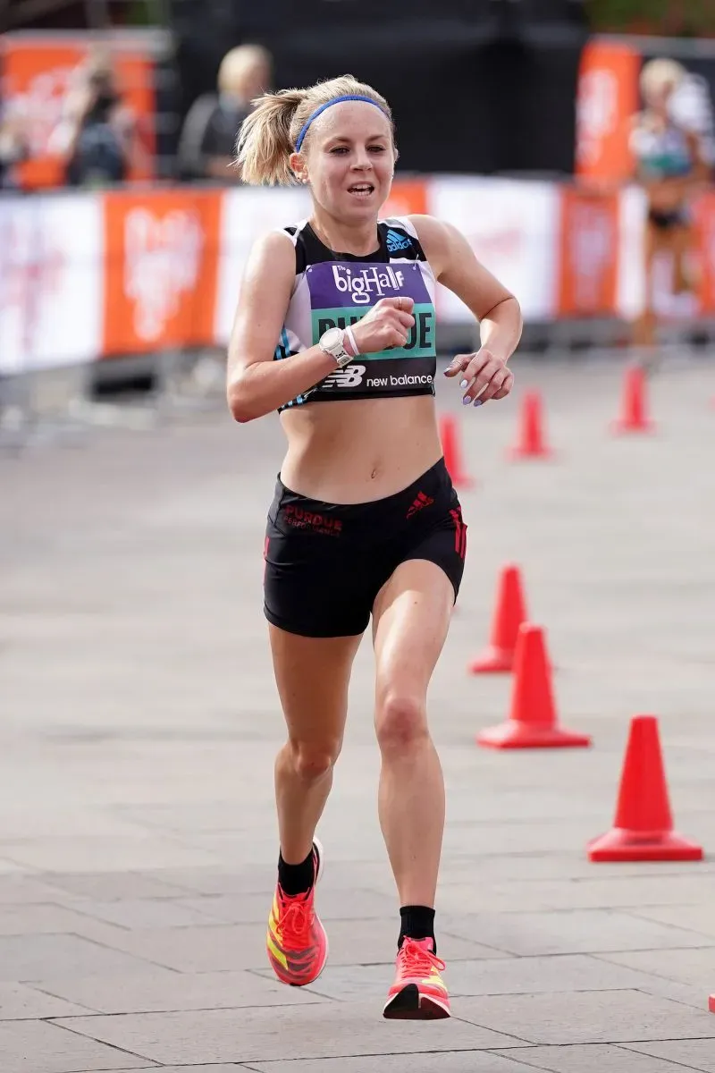 عداءة تجري في سباق، ترتدي ملابس رياضية سوداء وحذاءً رياضيًا ملونًا، مع خلفية تضم حواجز مرورية، تعكس أهمية التجميل والثقة في الأداء الرياضي.