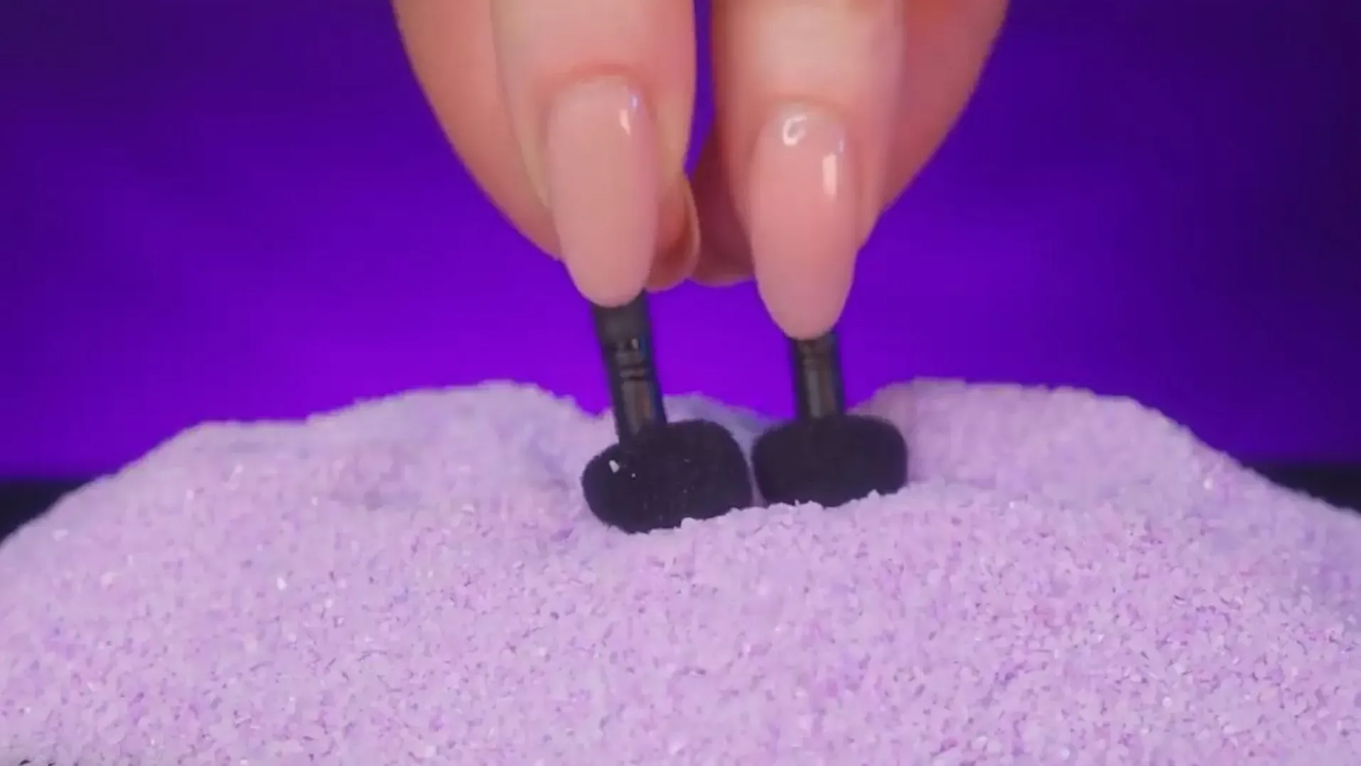 أصابع تضع سماعات أذن سوداء في رمال بنفسجية، مما يشير إلى أهمية الاستماع للموسيقى أو الكتب الصوتية كوسيلة للمساعدة على النوم.