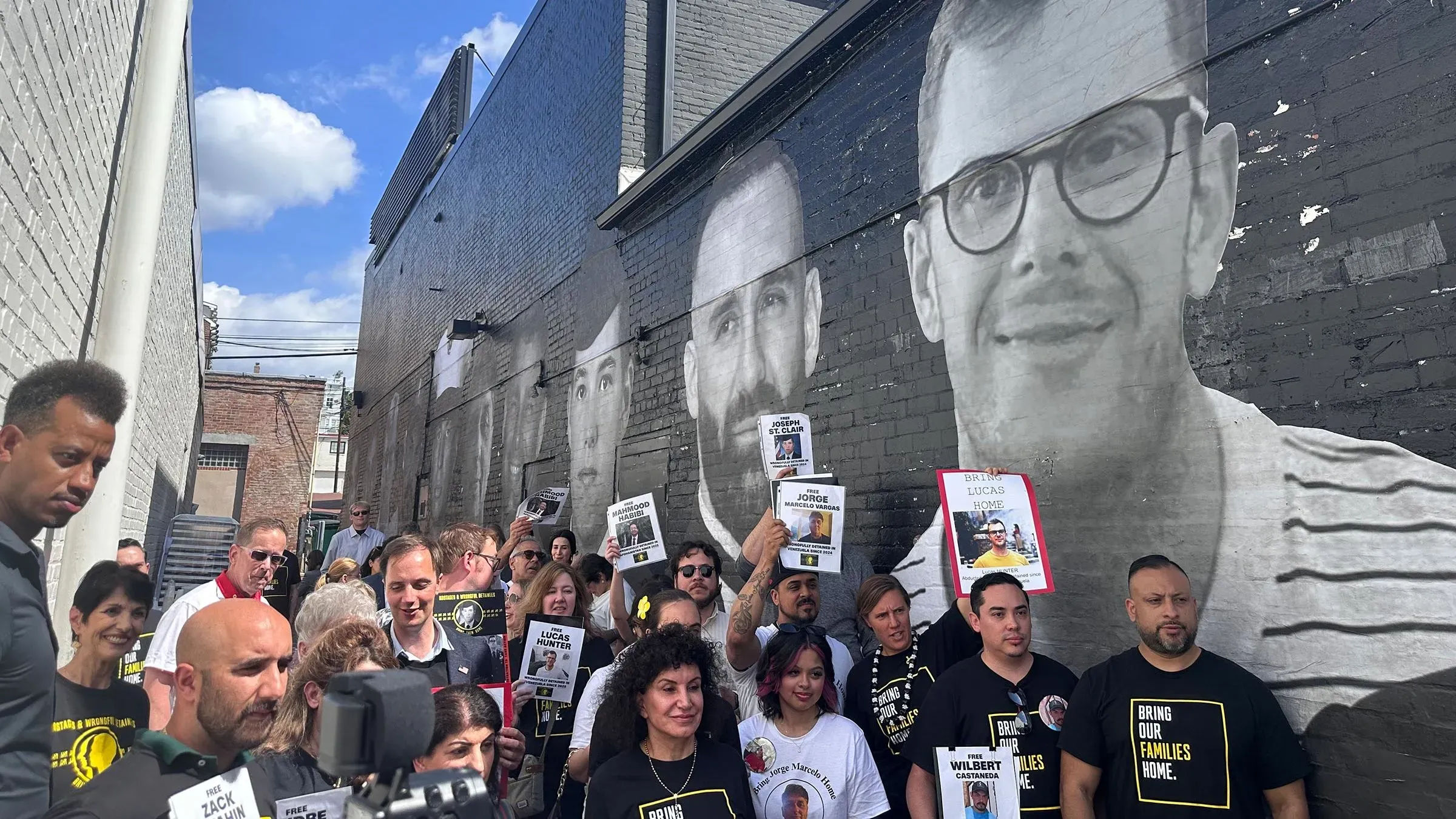 مجموعة من الأشخاص يتجمعون أمام لوحة جدارية ضخمة تعرض صور معتقلين أمريكيين في الخارج، يحملون لافتات للمطالبة بالإفراج عنهم.