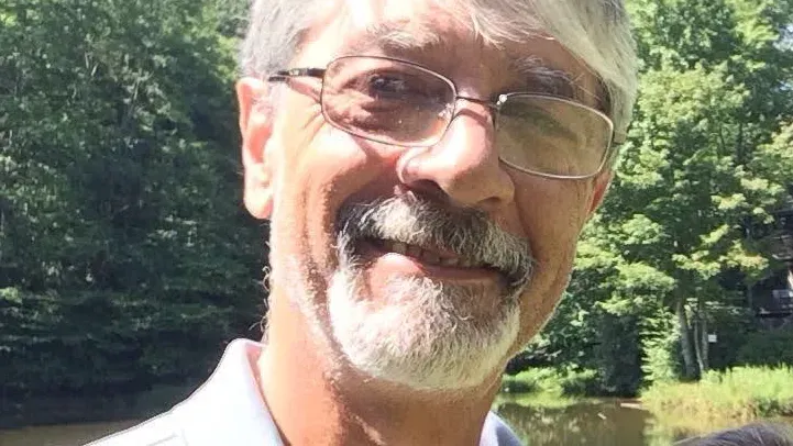 صورة لرجل مسن ذو لحية وشعر رمادي، يرتدي نظارات، مبتسم في بيئة طبيعية. تعكس الصورة معاناة محتجزين في مركز الهجرة في فلوريدا.