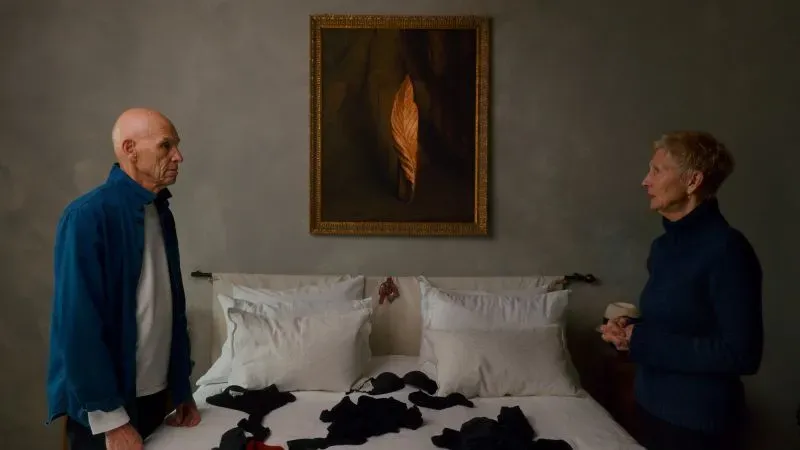 زوجان مسنّان يتحدثان في غرفة نوم، مع لوح فني على الجدار وملابس مطوية على السرير، يعكسان حميمية العلاقة بين الفنانين.