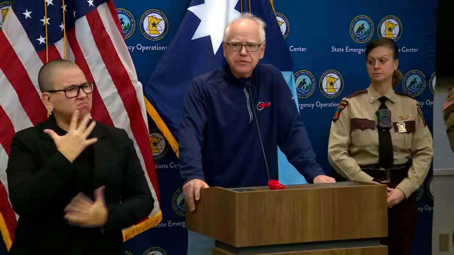 حاكم مينيسوتا تيم والز يتحدث في مؤتمر صحفي حول الوضع الأمني بعد حادث إطلاق النار، مع وجود أعلام خلفه وموظفين حكوميين.