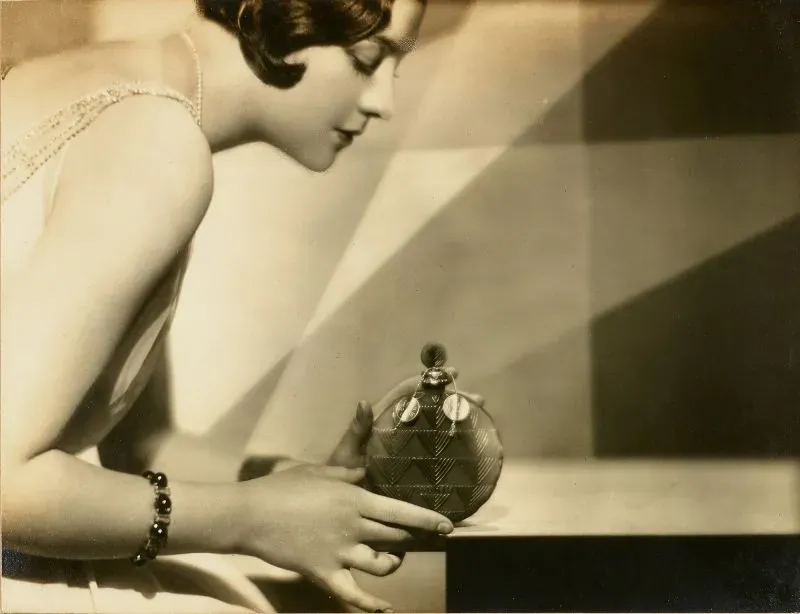 امرأة ترتدي فستانًا أنيقًا وتستعرض زجاجة عطر "Je Reviens" من تصميم تشارلز فريدريك وورث، في سياق معرض يبرز إرثه في الأزياء الراقية.